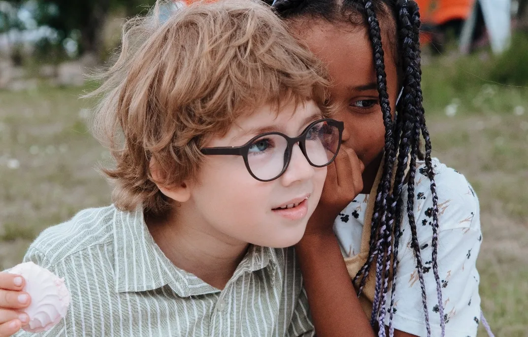 Ein Mädchen flüstert einem Jungen mit Brille ins Ohr, während er ein Gebäck hält.