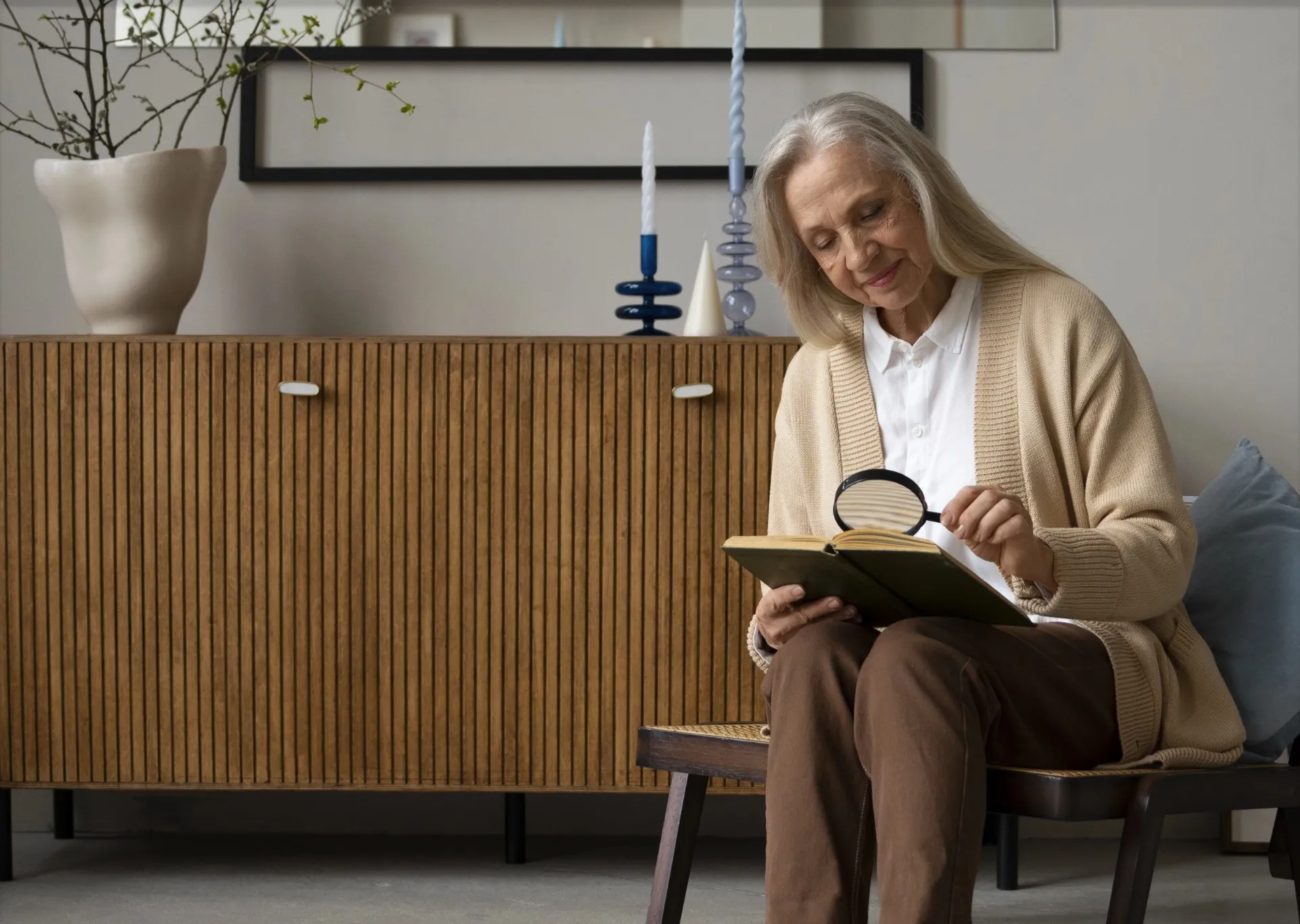 Ältere Frau sitzt auf einem Stuhl und liest ein Buch mit einer Lupe in einem modernen Wohnzimmer.