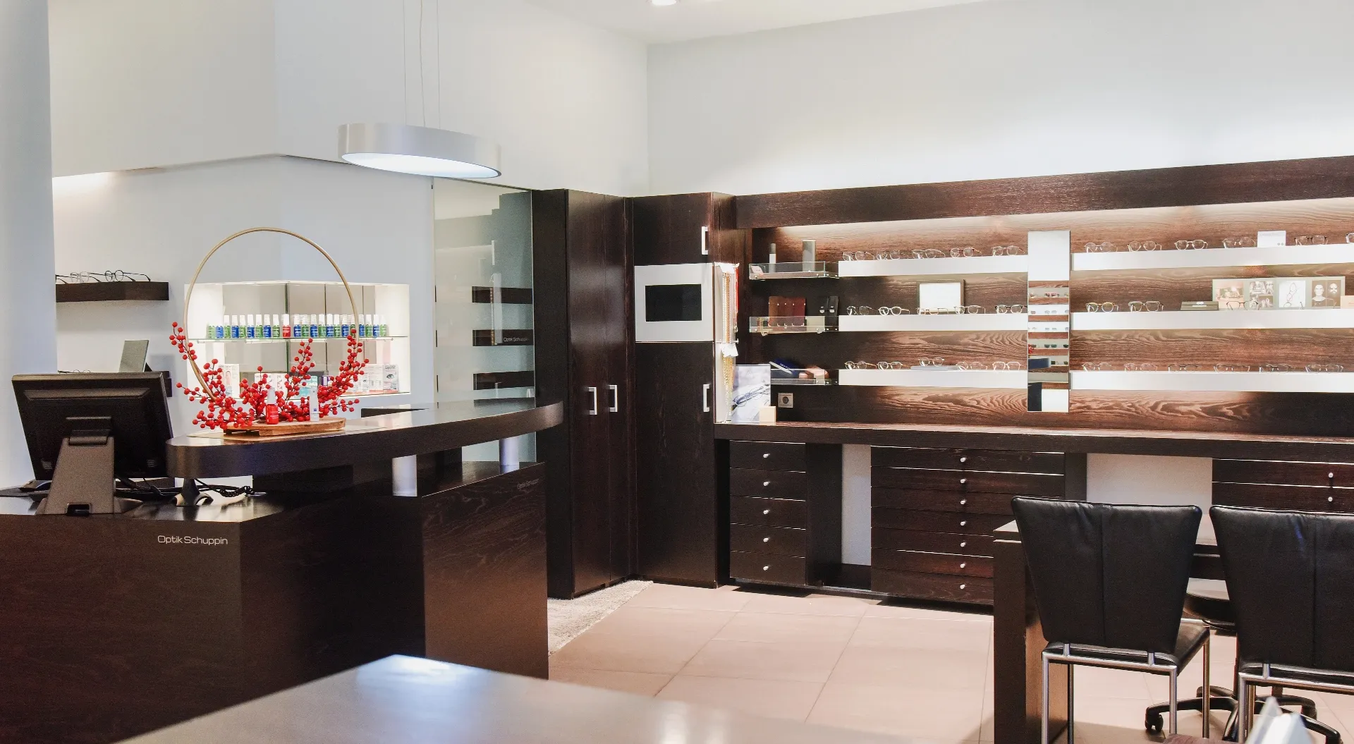Moderne Optiker-Filiale mit dunklen Holzregalen, einem Tresen mit roter Dekoration und Glasflaschen sowie zwei schwarzen Stühlen.