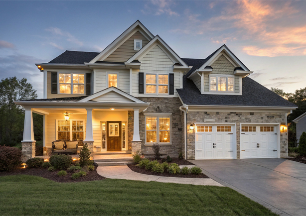 Large two-story house with illuminated windows, front porch with swing, double garage, and landscaped yard at sunset.