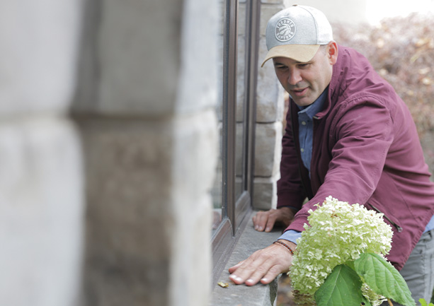 Man in a maroon jacket and gray cap inspecting a window ledge outside a stone building.