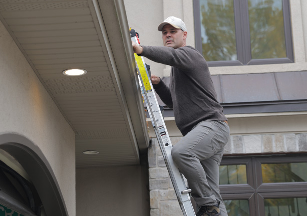 Man in a gray sweater and cap climbing a ladder to inspect the roof edge of a house.