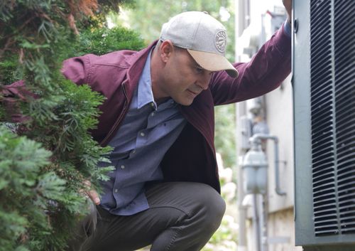 Man wearing a cap and jacket inspecting an outdoor air conditioning unit next to a house.