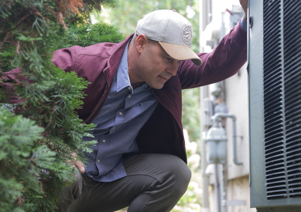 Man wearing a cap and jacket inspecting an outdoor air conditioning unit next to a house.