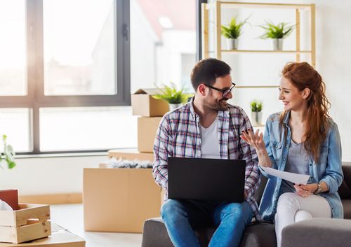 Smiling couple sitting on a couch with a laptop and documents, surrounded by moving boxes in a bright room.
