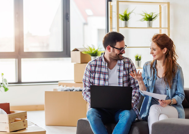 Smiling couple sitting on a couch with a laptop and documents, surrounded by moving boxes in a bright room.