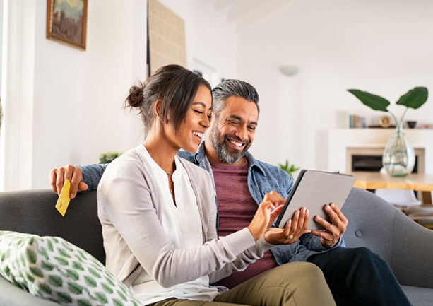 Smiling couple sitting on a couch using a tablet, with the woman holding the device and the man holding a credit card.