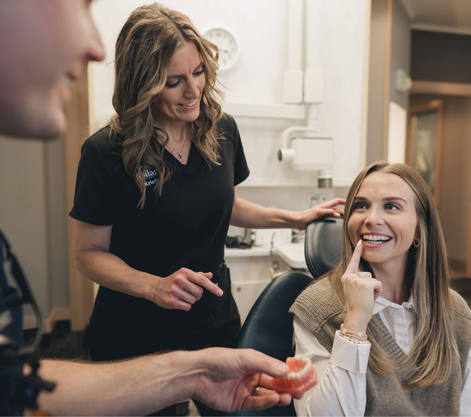 Dentist showing a dental model to a woman sitting in dental chair who is smiling and pointing to her teeth.
