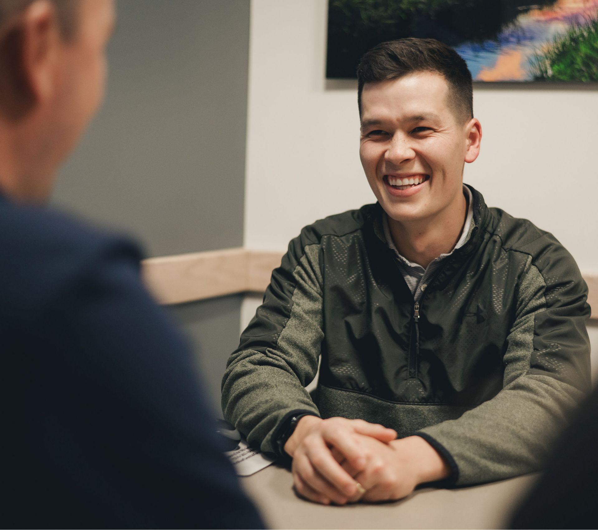 Smiling young man in a green jacket sitting at a table and engaging in conversation.