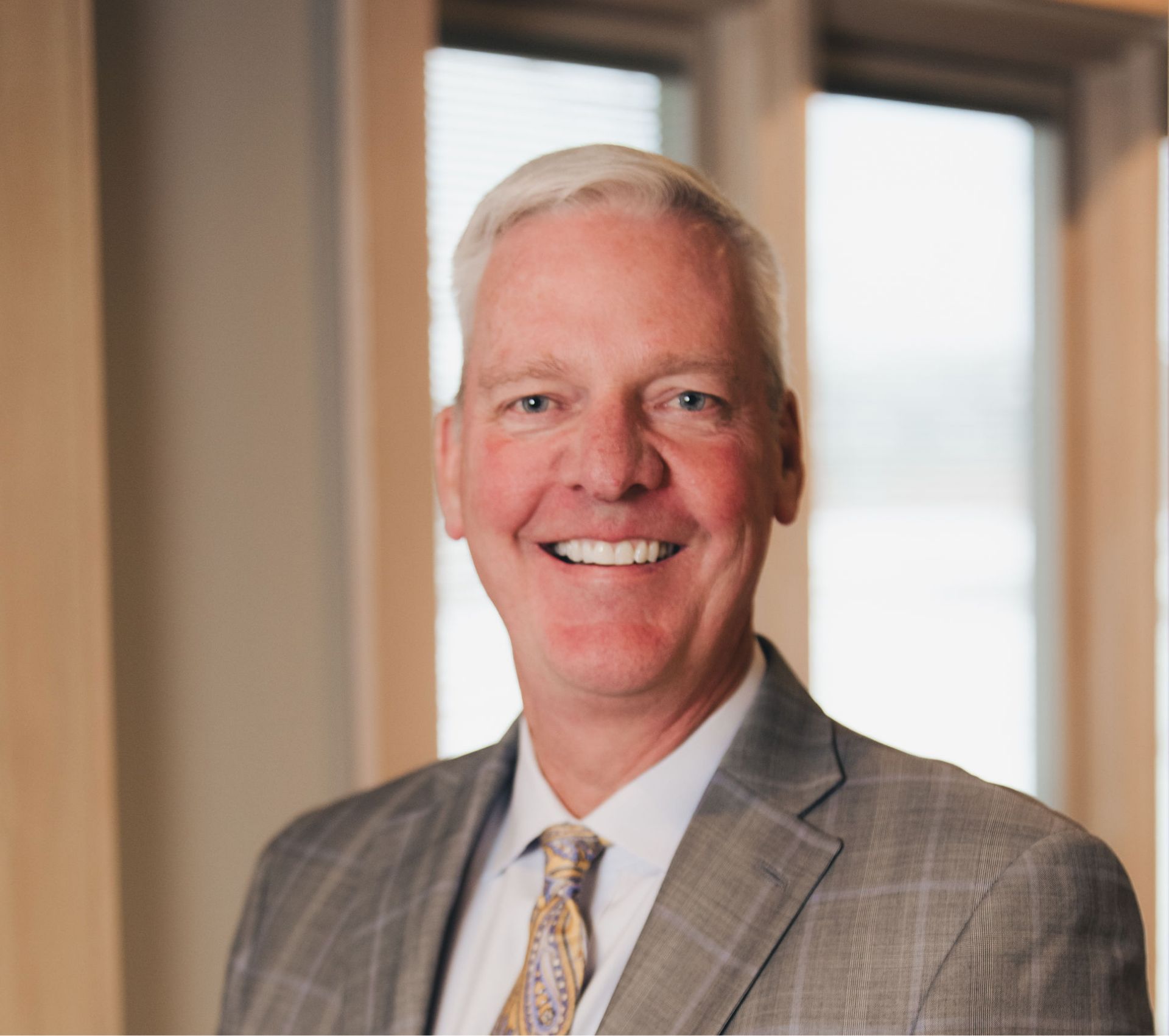 Smiling middle-aged man with short white hair wearing a gray plaid suit and paisley tie in an office setting.