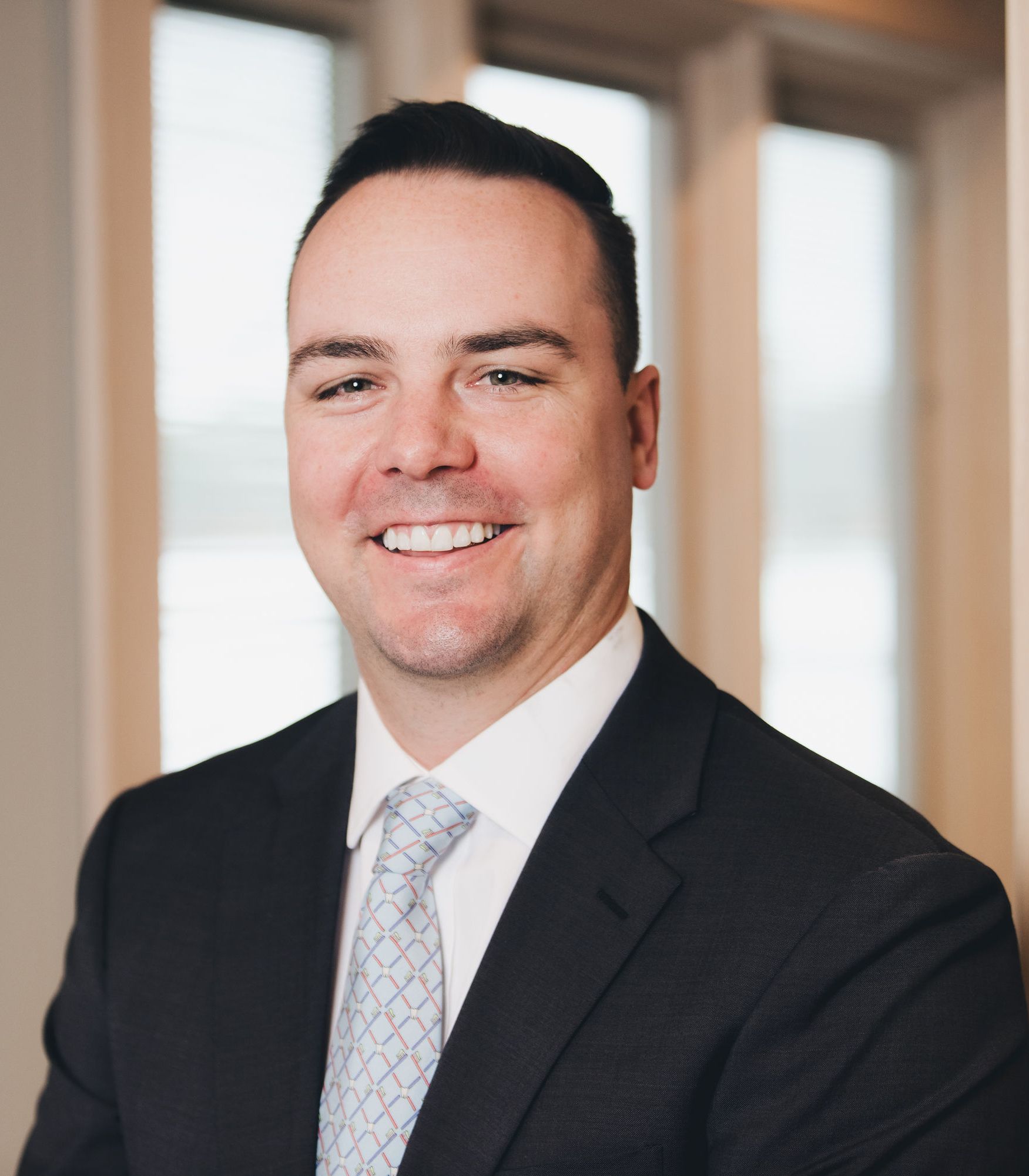 Smiling man in a dark suit and patterned tie standing indoors with blurred windows in the background.