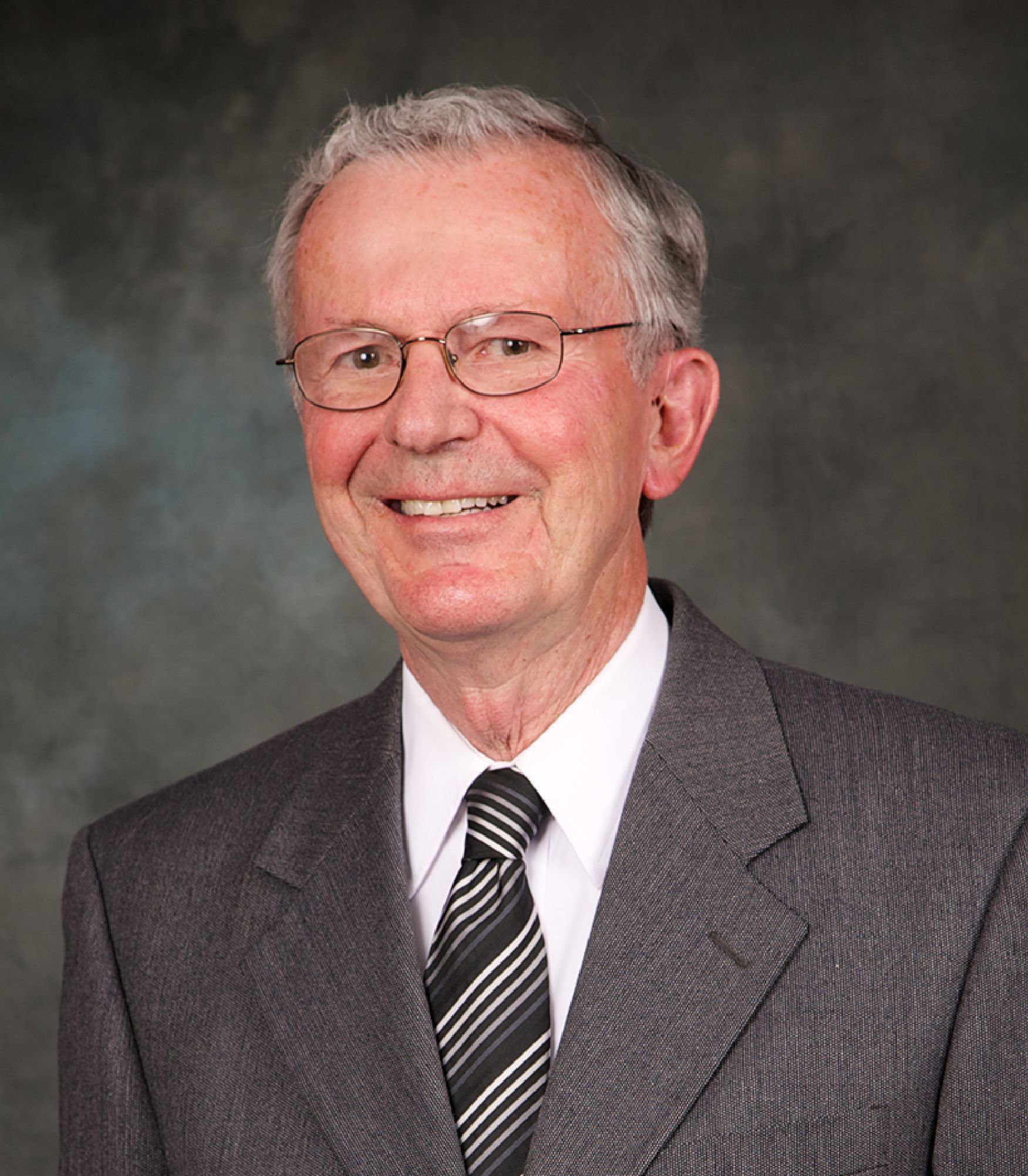 Smiling older man with gray hair and glasses wearing a gray suit, white shirt, and striped tie against a dark background.