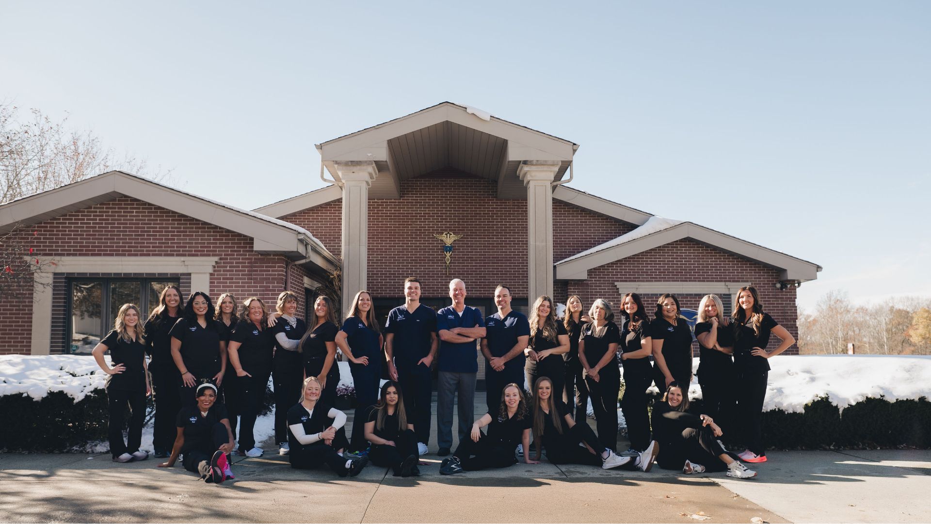 A large group of diverse medical staff in black scrubs posing outside a brick building with snow on the ground.