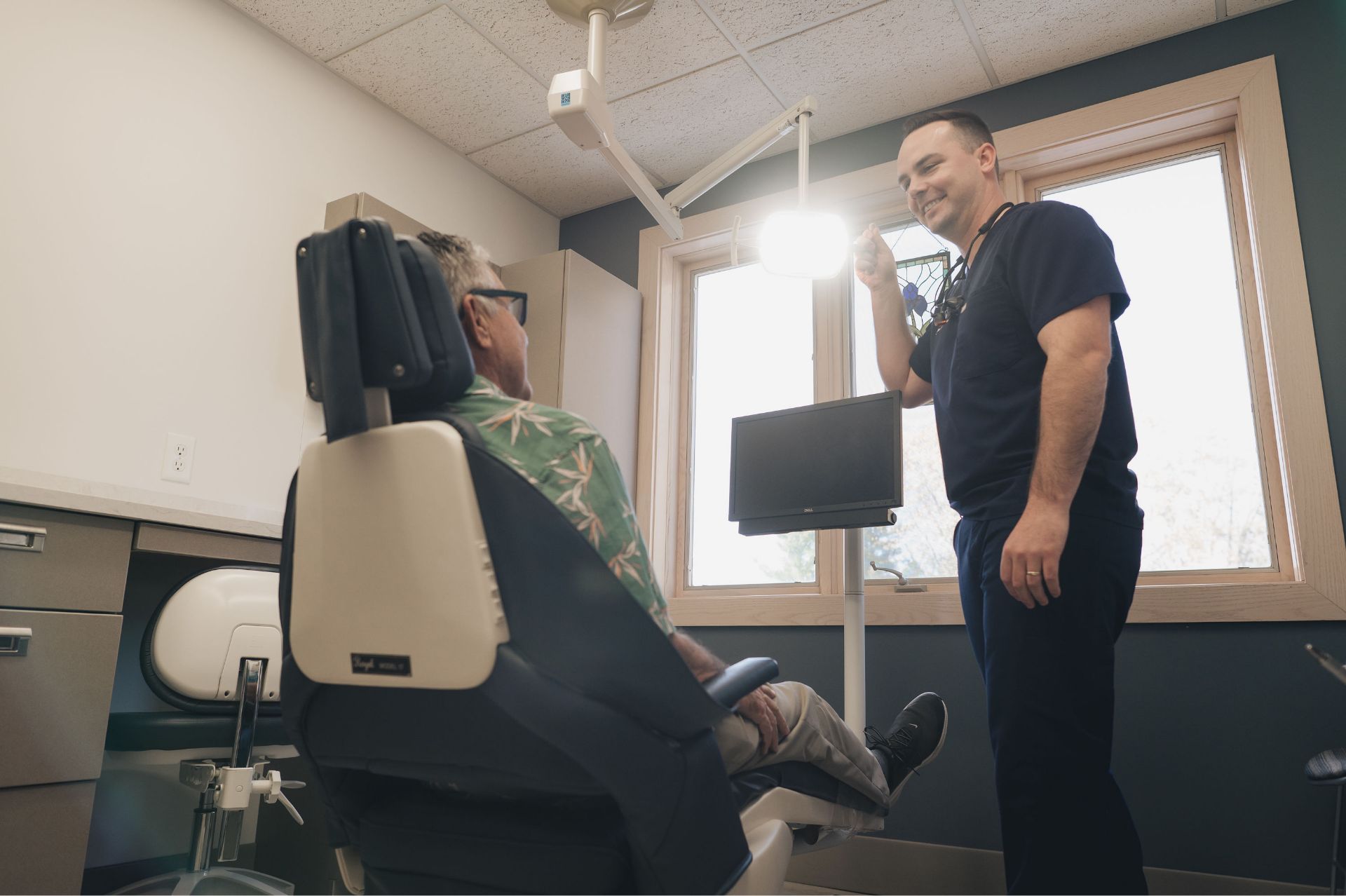 Dentist in navy scrubs stands next to dental chair talking to a seated patient in a dental office.