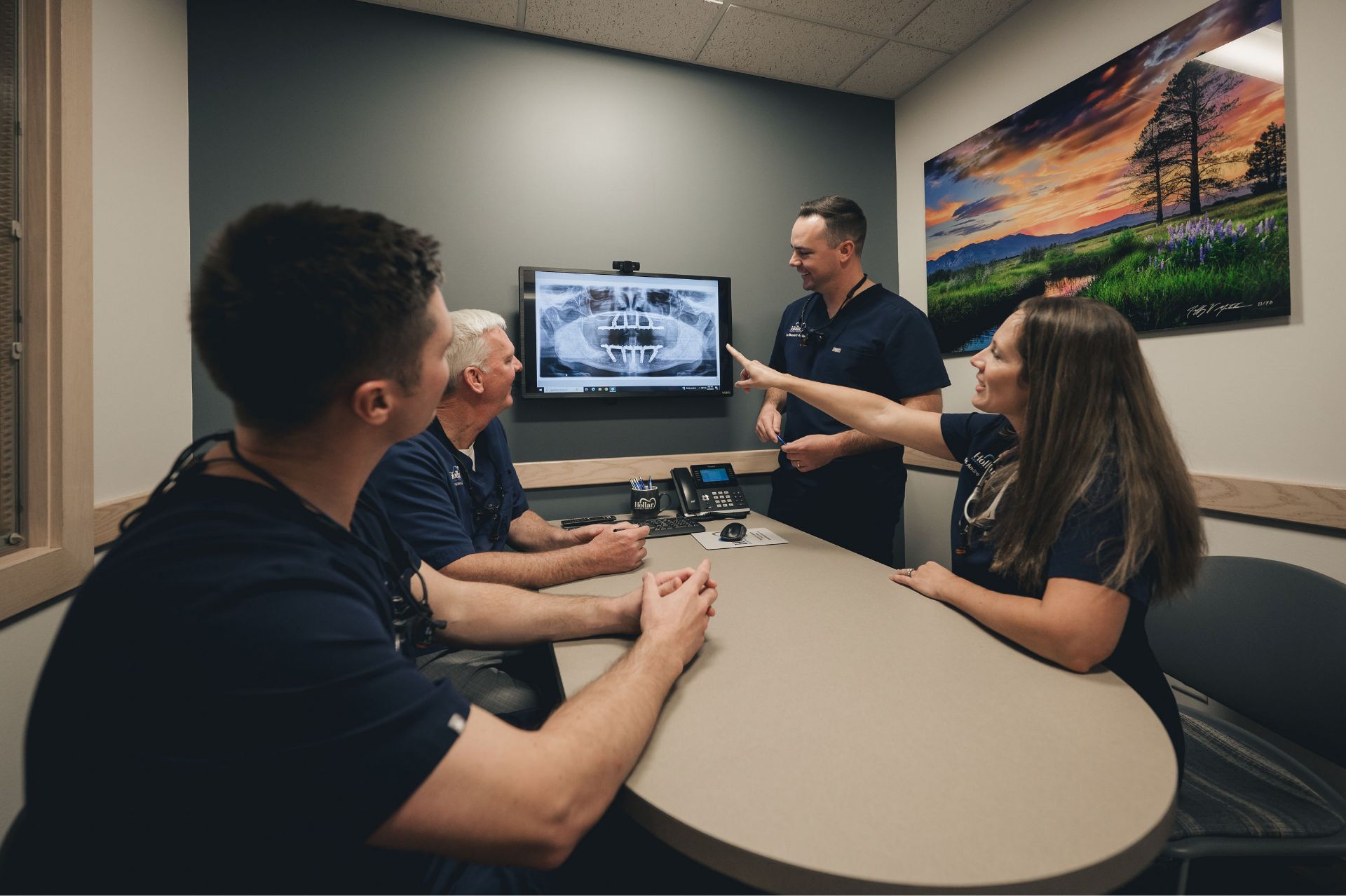 Four dental professionals in navy scrubs reviewing a dental X-ray displayed on a wall-mounted screen in a conference room.