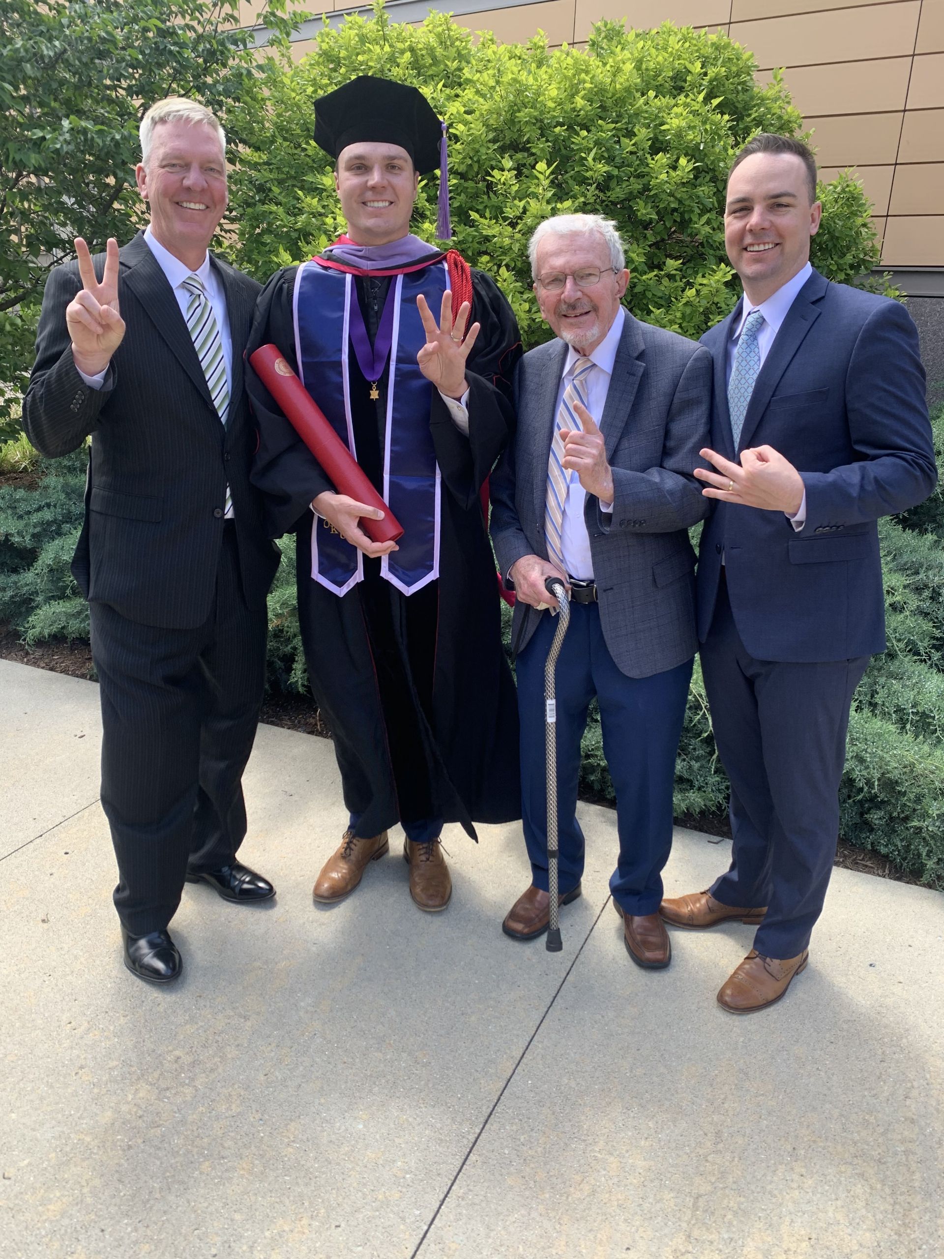 Four men standing outside with greenery in the background; one is wearing a graduation gown and cap holding a diploma, while the others wear suits and make hand signs.