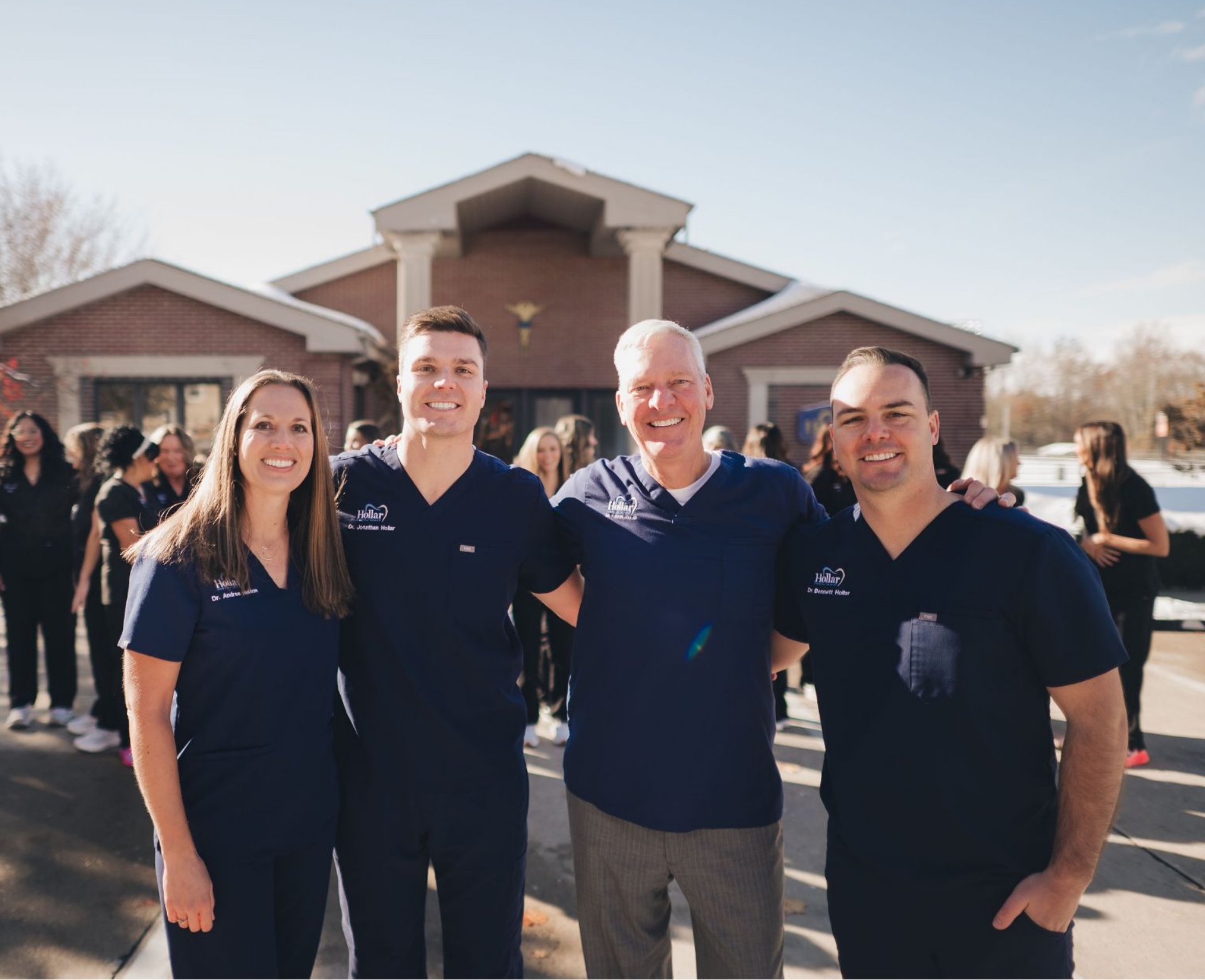 Four medical professionals in navy scrubs smiling outdoors in front of a brick building with a group of colleagues in the background.