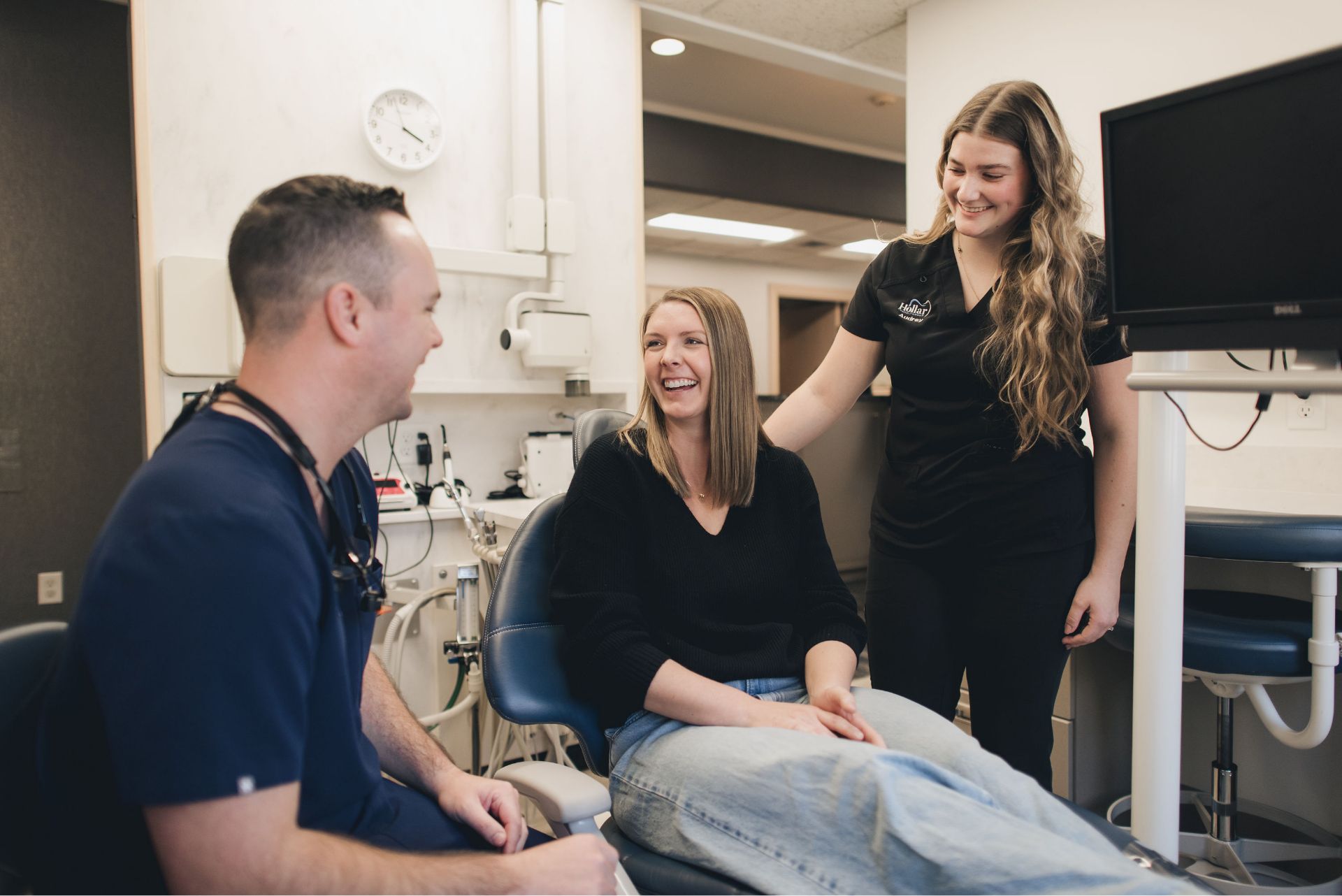 A female patient smiling and sitting in a dental chair while talking with a male dentist and a female dental assistant in a dental office.