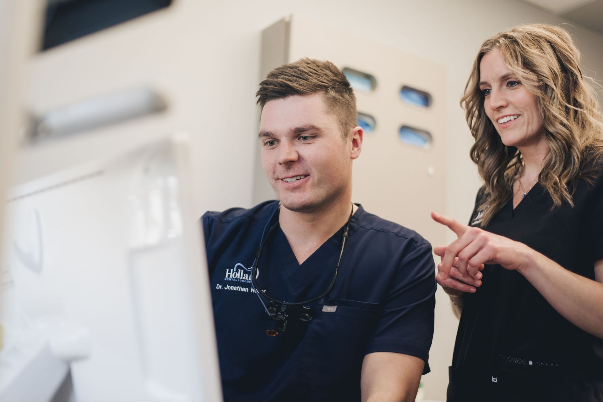Dental professional wearing navy scrubs with magnifying glasses looks at a screen while a woman in black scrubs points and smiles beside him.