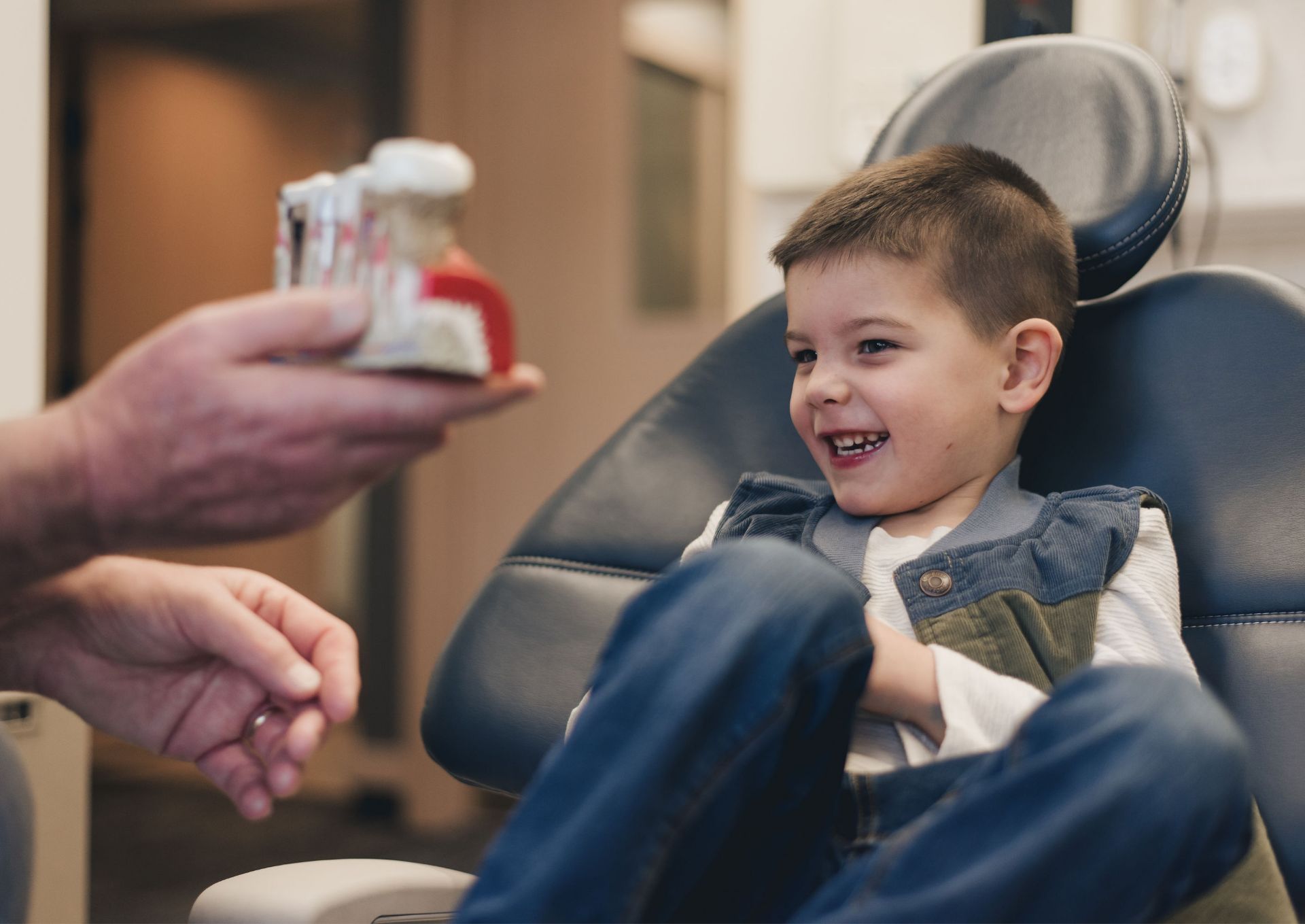 Young boy sitting in a dental chair smiling while a person shows a dental model.