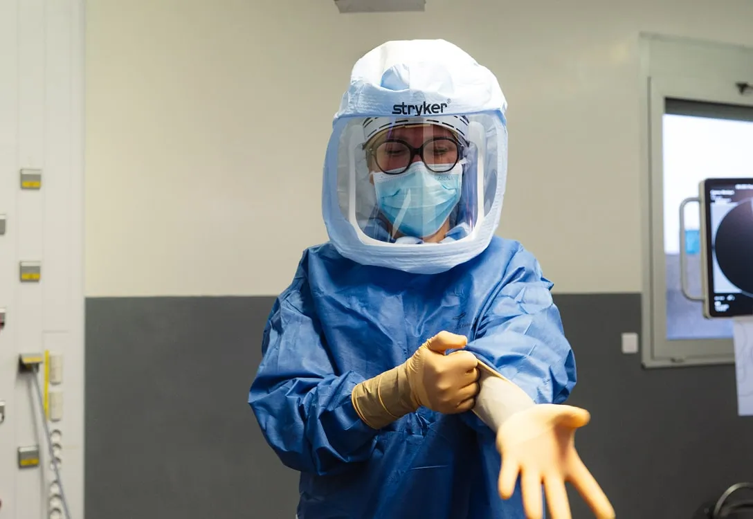 Professionnel de santé portant une combinaison de protection complète, un masque et un casque avec visière, enfilant des gants en latex dans un environnement médical.
