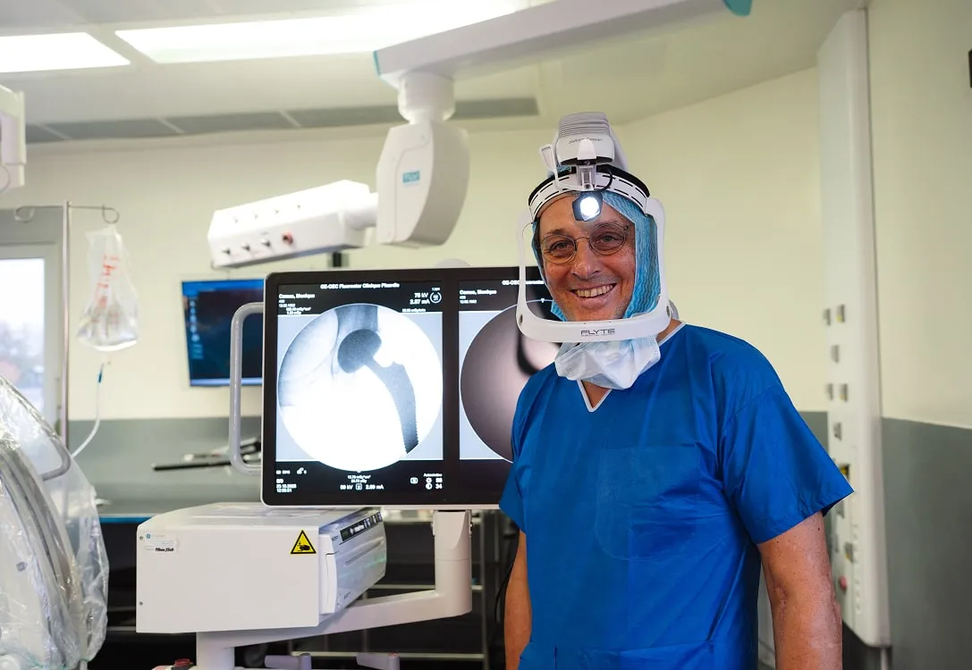 Chirurgien souriant en tenue bleue avec casque de protection lumineux dans une salle d'opération moderne devant un écran montrant une radiographie d'implant orthopédique.