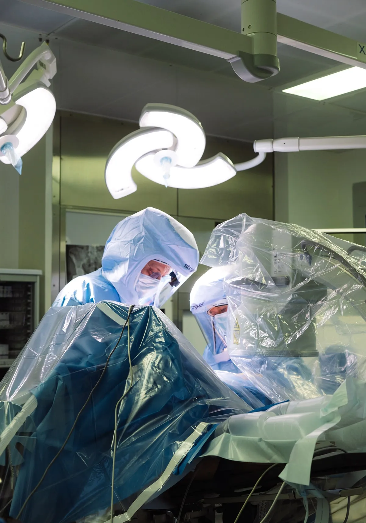 Deux chirurgiens en tenue de protection complète effectuent une opération sous des lampes chirurgicales dans une salle d'opération stérile.