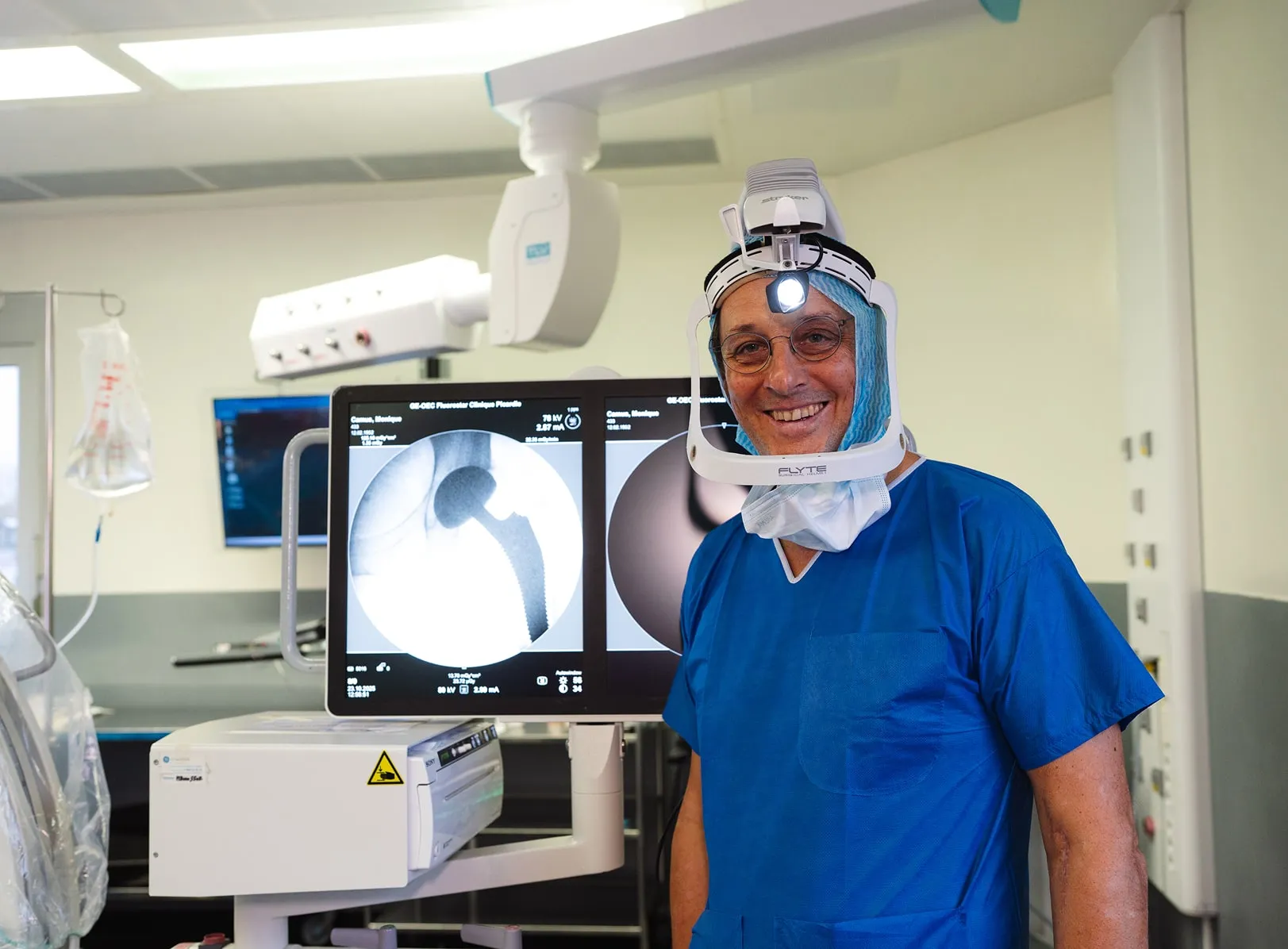 Chirurgien souriant en tenue bleue avec casque à lumière dans une salle d'opération devant un écran montrant un implant de hanche.