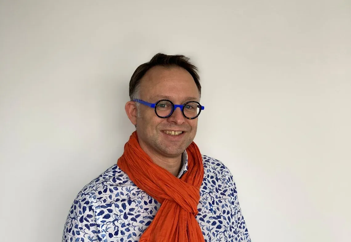 Homme souriant portant des lunettes rondes bleues, une chemise à motifs floraux bleus et une écharpe orange.