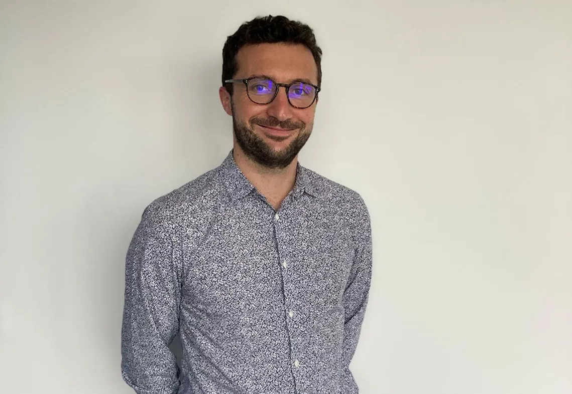 Homme portant des lunettes et une chemise à motifs, souriant devant un mur blanc.