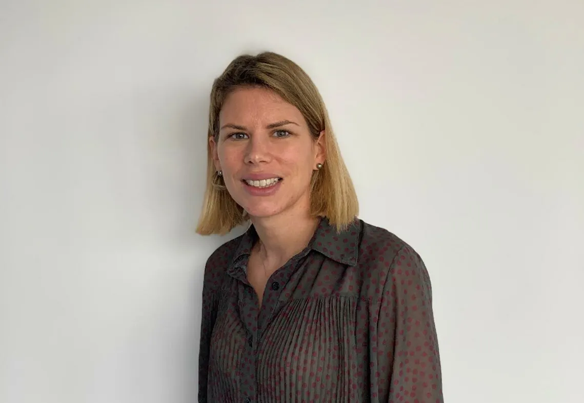 Portrait d'une femme souriante aux cheveux blonds mi-longs, portant une blouse sombre à pois rouges, devant un mur blanc.
