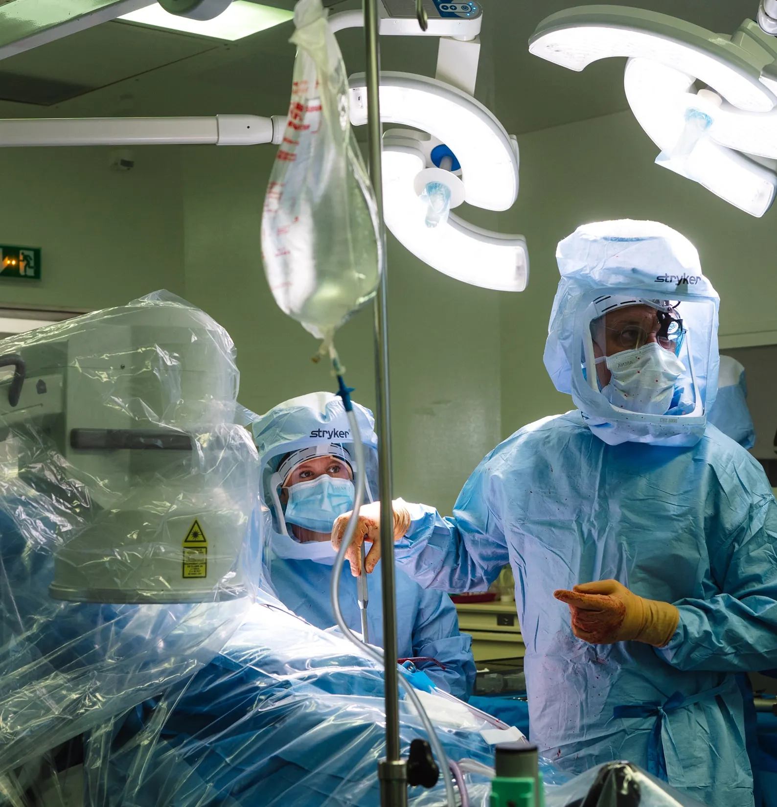 Deux chirurgiens en tenue stérile avec masques et protections faciales travaillent dans une salle d'opération sous des lampes chirurgicales.