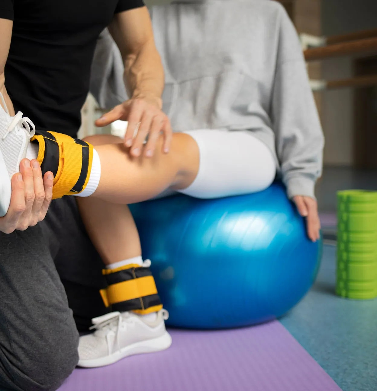 Personne assise sur un ballon de gym bleu effectuant des exercices avec des poids aux chevilles sous la supervision d'un entraîneur.