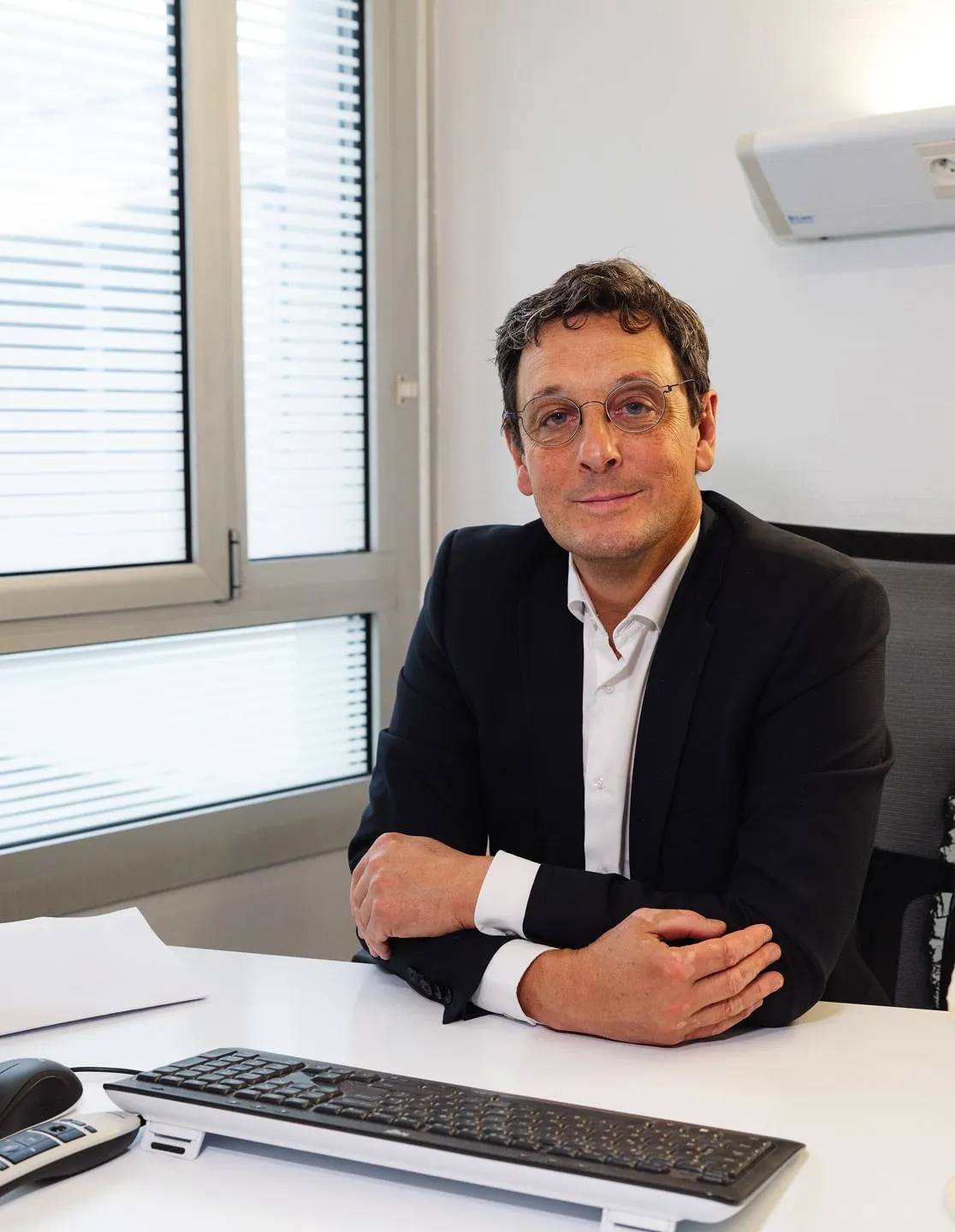 Homme portant des lunettes et un costume noir assis à un bureau blanc avec un clavier et une souris devant lui.