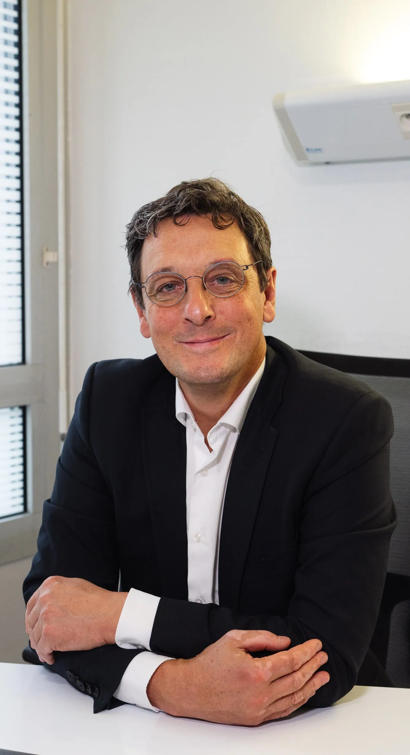Homme portant des lunettes, veste noire et chemise blanche, assis à un bureau avec les bras croisés et souriant légèrement.