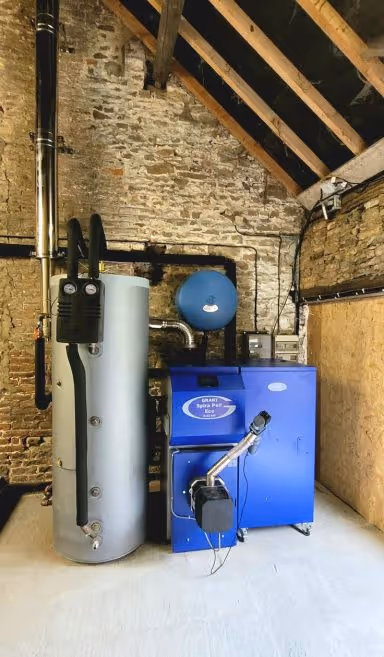 Chaudière bleue avec réservoir d'eau gris installée dans une pièce avec murs en pierres apparentes et plafond en poutres de bois.