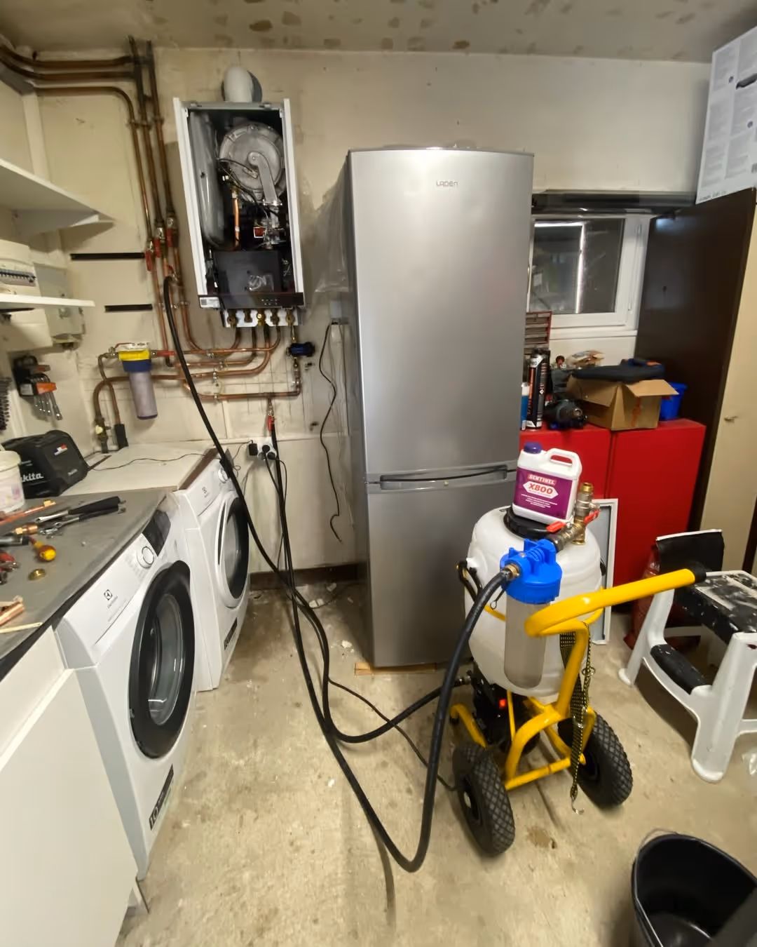 Salle de buanderie avec chaudière murale ouverte, réfrigérateur argenté, lave-linge, sèche-linge et équipement de maintenance sur un chariot jaune.