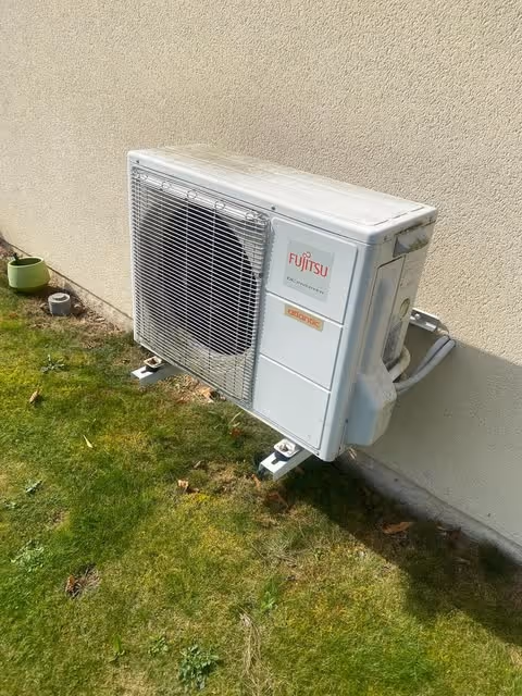 Unité extérieure d'une pompe à chaleur Fujitsu montée sur un mur beige au-dessus d'une pelouse verte.