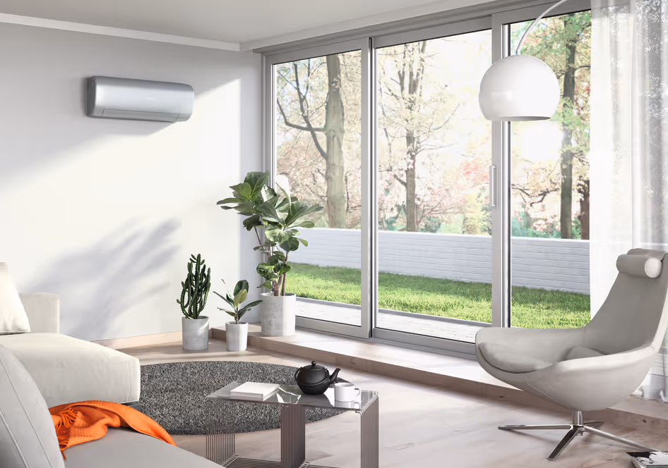 Salon moderne avec fauteuil gris, canapé, table basse en verre, plantes en pots, et grandes baies vitrées donnant sur un jardin.