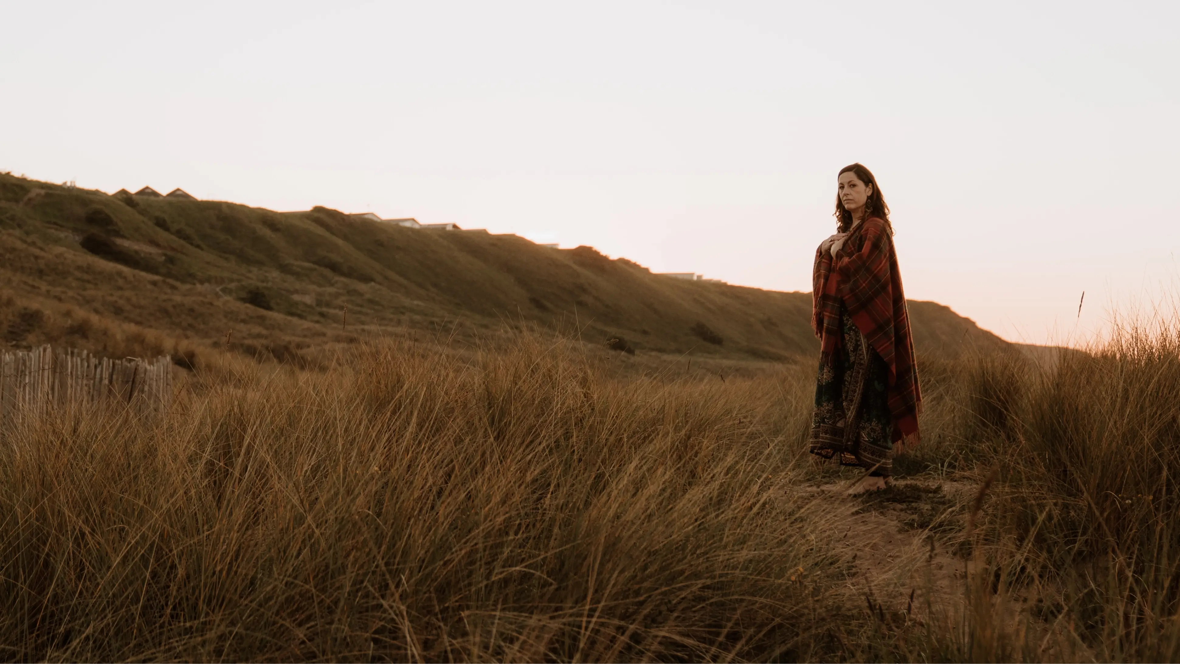 Dawn wrapped in a red plaid shawl standing barefoot in tall grass facing the camera at sunset.