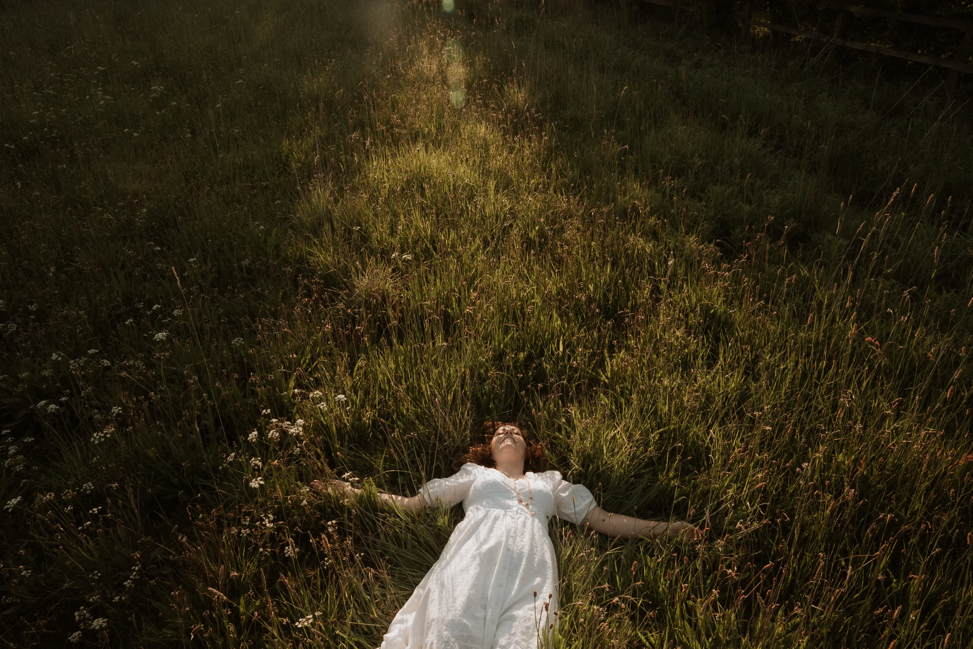 Dawn in white dress lying on grass in a sunlit meadow with arms outstretched and eyes closed.