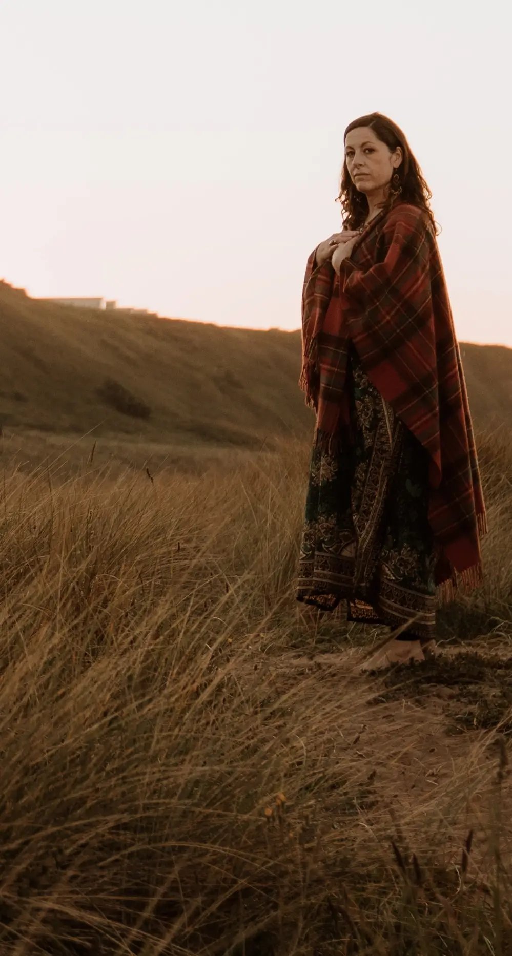 Dawn wrapped in a red plaid shawl standing barefoot in tall grass facing the camera at sunset.
