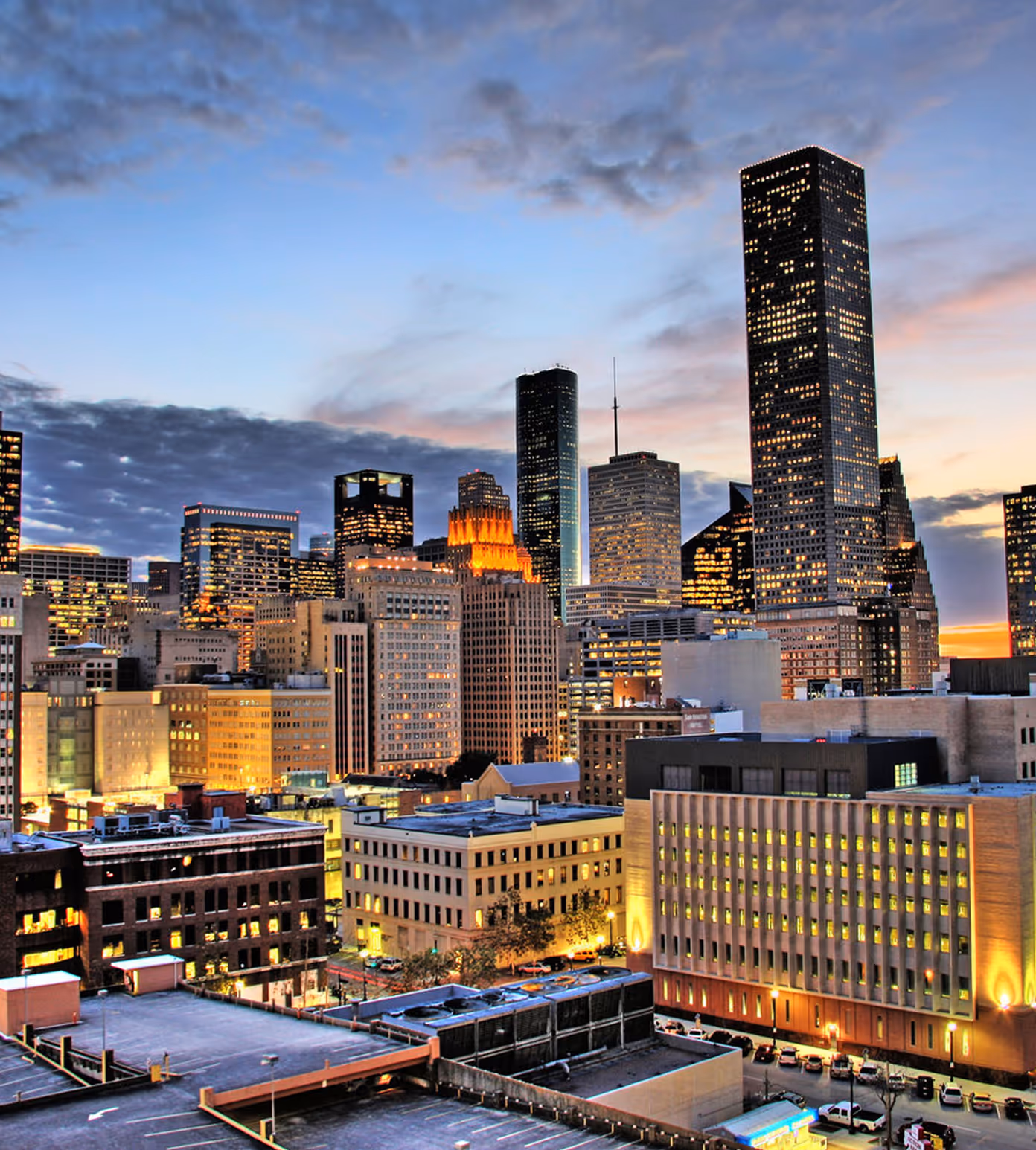 City skyline at dusk with tall buildings illuminated against a partly cloudy sky.