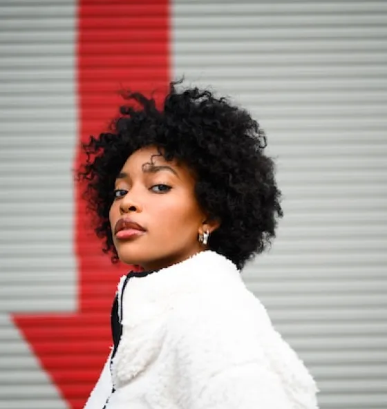 Person with curly hair wearing a white coat standing in front of a red arrow on a gray wall.