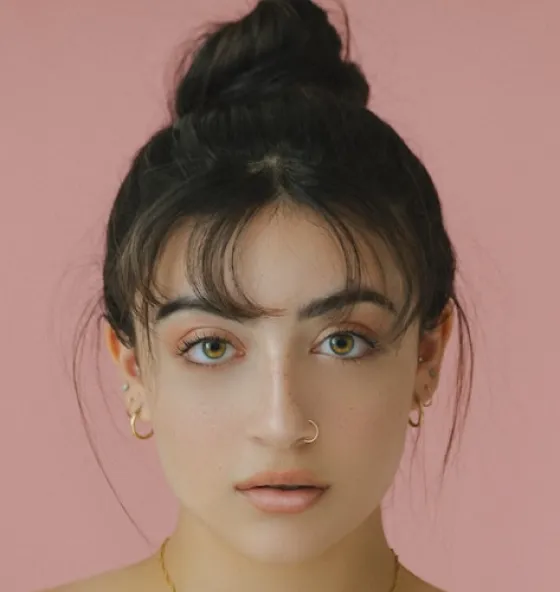 Person with hair in a bun wearing gold earrings against a pink background.