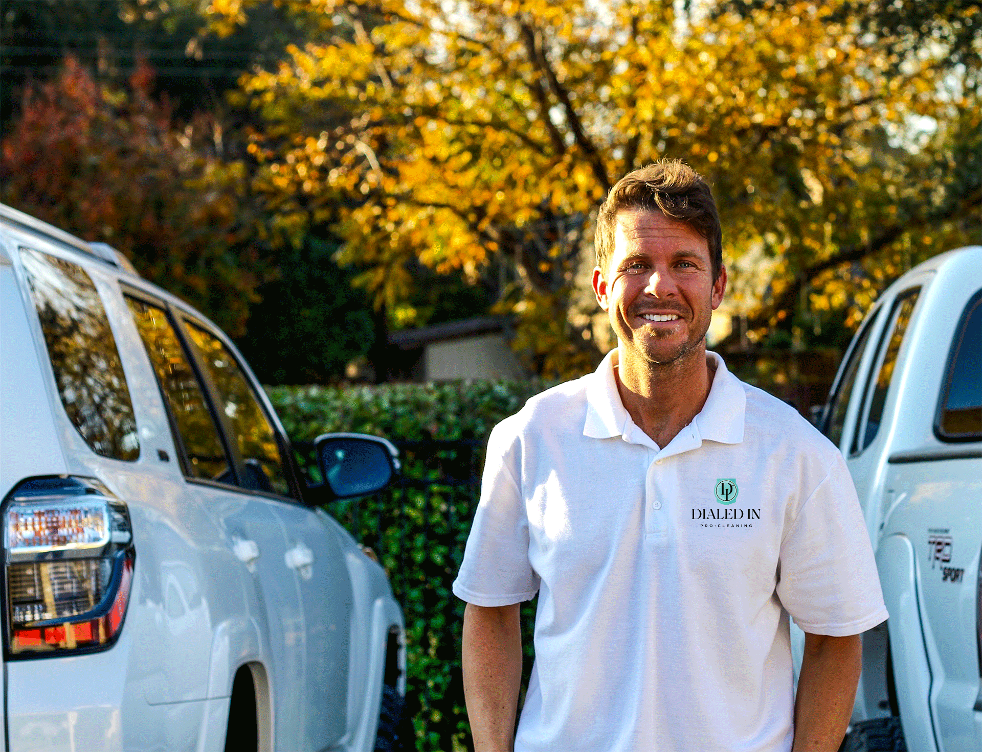 Smiling man wearing a white Dialed In Pro-Cleaning polo shirt stands between two white vehicles outdoors with autumn trees in the background.