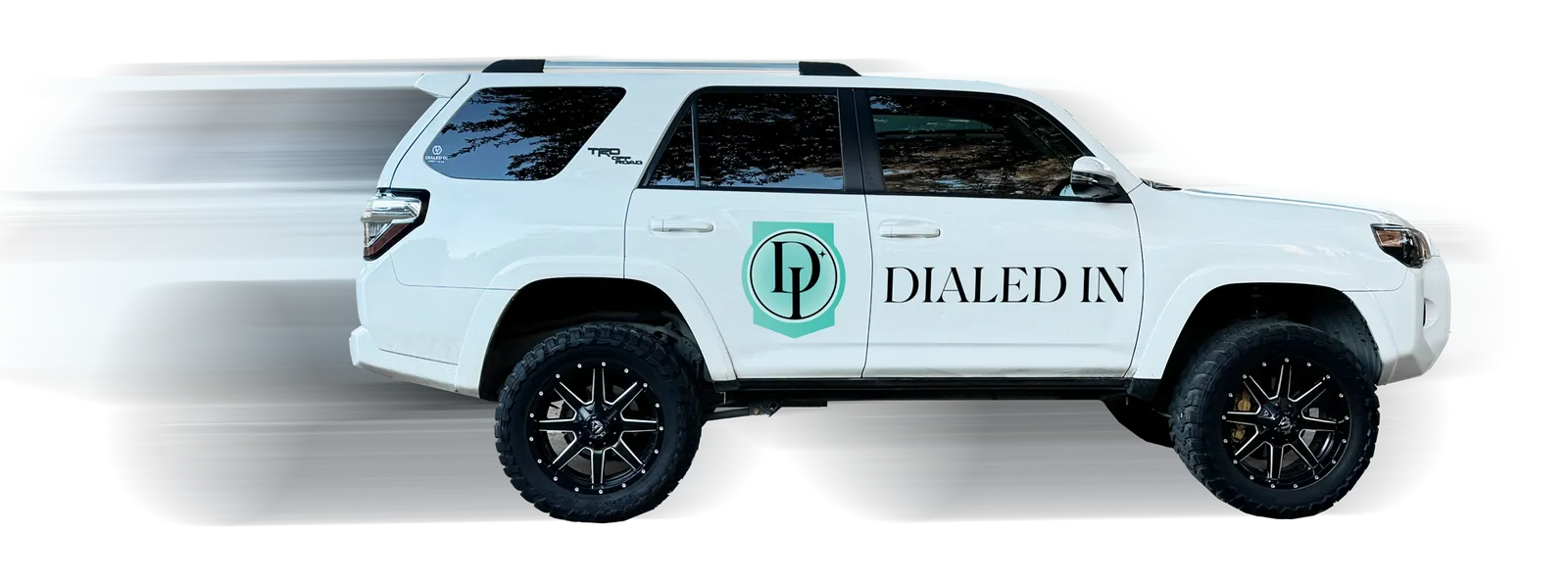 White SUV with black wheels featuring 'Dialed In' logo and text on the side, motion blur behind the vehicle.