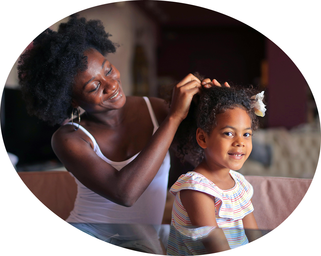 A woman in a white tank top gently styles a young girl's hair. The girl, in a striped shirt, smiles warmly, creating a tender, loving atmosphere.