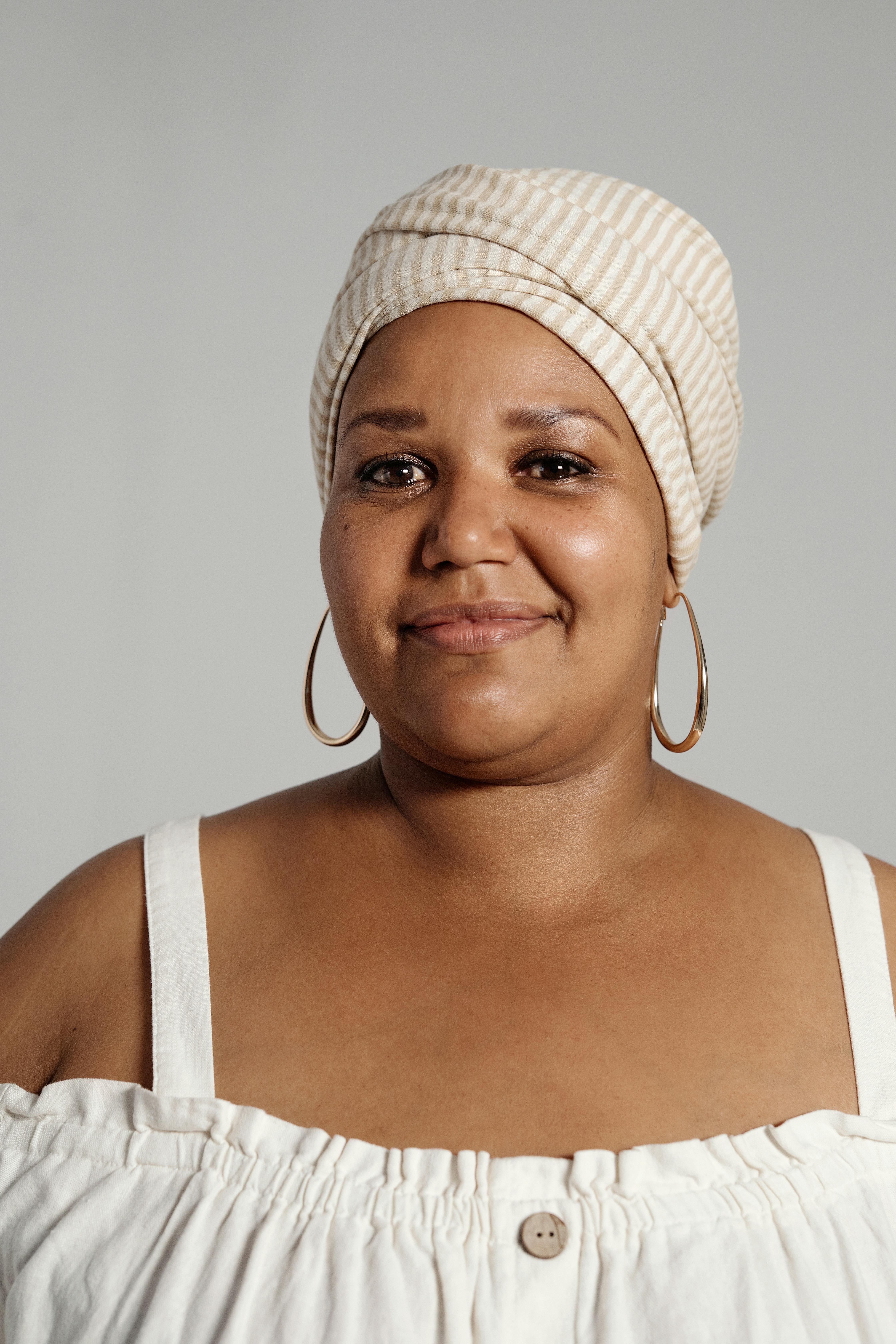 A woman wearing a striped head wrap and hoop earrings smiles softly. She is dressed in a white off-shoulder top. The background is plain and light gray.