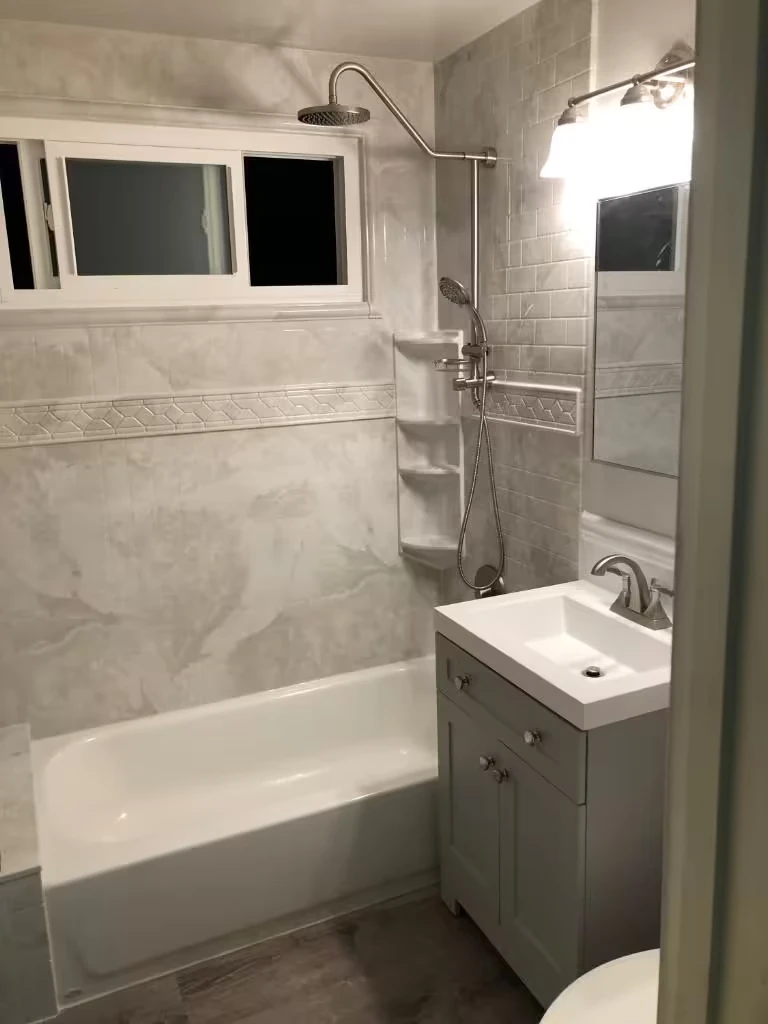 Modern bathroom with gray tiled bathtub and shower, white toilet with rolled towel, and vanity with sink and green leaf in vase.